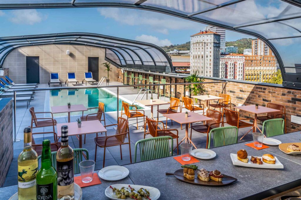A rooftop terrace at Axel Bilbao with finger food laid out on the bar. Behind is a pool and views of the city.