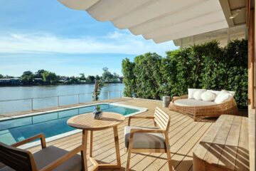 Views from a riverside suite terrace. Blonde wood decking and outdoor furniture with a blue pool against its edge, leading out to a still river. Sala Bang Pa-In is one of the most romantic getaways near Bangkok.