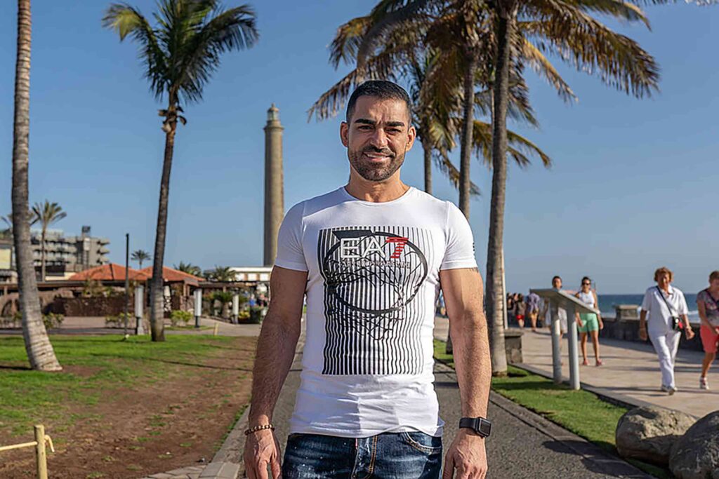 Fernando Ilarduya, a muscly Spanish man with short brown hair, thichk eyebrowns, moustache and beard, stands in front of the Faro lighthouse in Meloneras beachfront. He wears a white EA7 tshirt and blue jeans