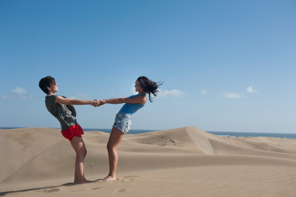 Two women, a couple, one with short brown hair wearing a grey vest and red shorts, the other with long brown hair, blue vest and cut-off jeans hold hands and lean away from each other to the backdrop of the sand dunes in Maspalomas, Gran Canaria