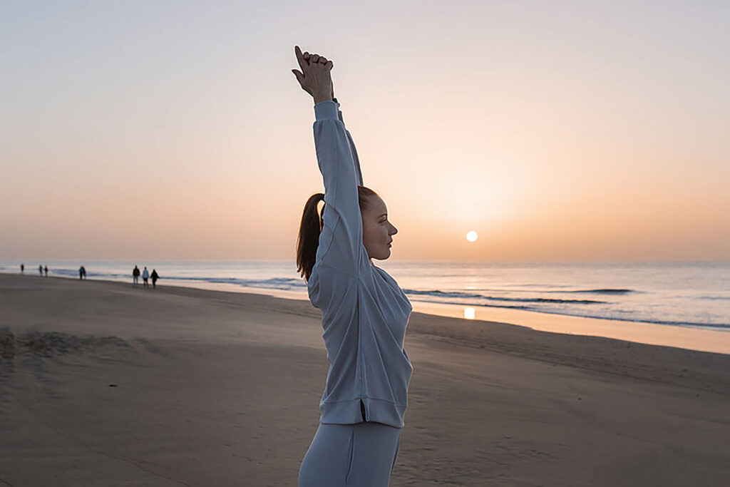 Eva Lopez with long brown hair tied into a pony tail looks out to sea as the sun rises and holds a sun salutation yoga pose. She is caucasian and wearing a fitted grey track suit.