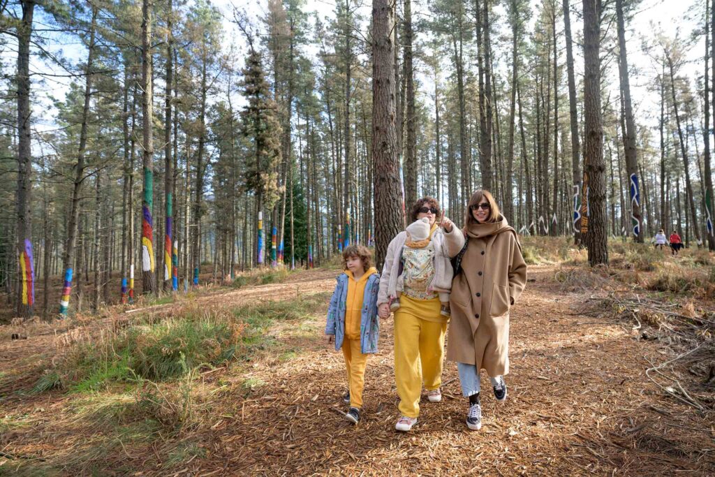 A lesbian couple and their two children walk through a forest with art installations on the trees.