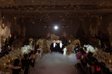Two Asian men in tuxedos walk way from a white grand piano in a puff of smoke, hand in hand as wedding guests watch and cheer in a beautifully decorated room. LGBTQ+ weddings at Capella Bangkok.