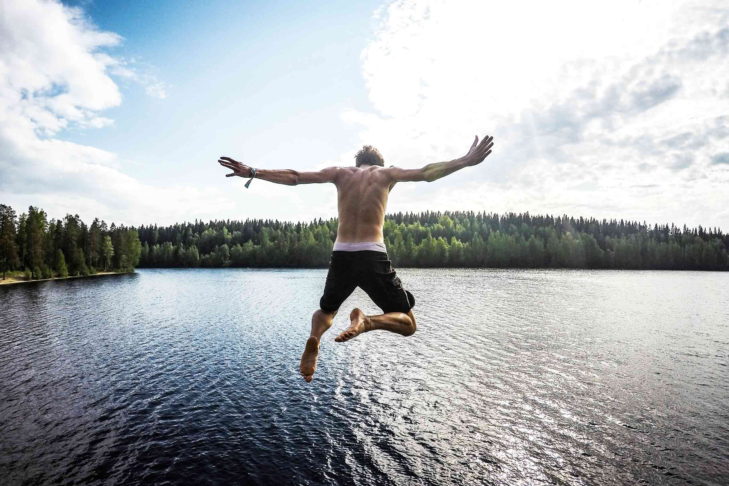 Topless man with long black shorts and pink underwear jump dives into the sea water in the archipelago around Stockholm. The background is a horizon of pine trees.