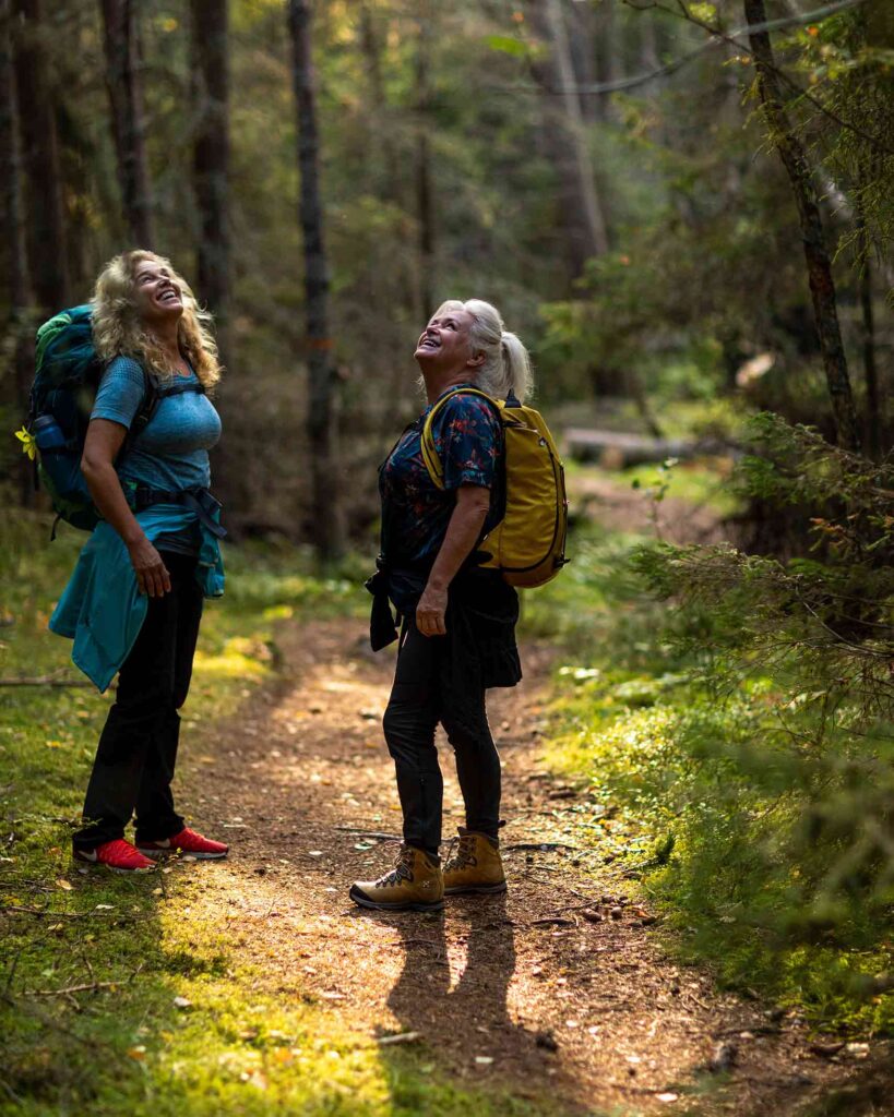 Two women, one with long blond hair, a blue t-shirt, backpack, black trousers and red shoes; the other with white hair, a flowery shirt, yellow backpack and tan shoes, look up into a beam of light in a forest and smile