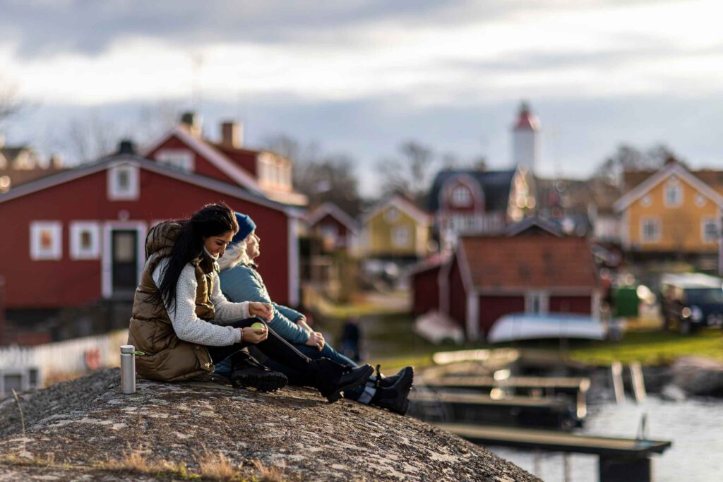 Two women in hiking great, one with long brown hair, the other in a blue hat sit in front of a waterside town in the Stockholm Archipelago. There are traditional Swedish buildings in the background, out of focus