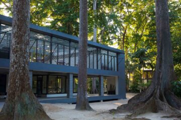A square blue building surrounded by forest. The downstairs has no walls and the upstairs has floor-to-ceiling windows.