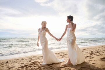 Two women walk hand in hand down a beach in Thailand in wedding dresses. Both are white, one has long blonde hair, the other short dark hair
