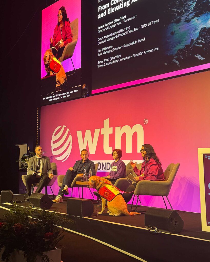 Four panelists seated on stage with a large logo behind them. One panellist speaks into a microphone while a guide dog in a red vest sits at their feet.