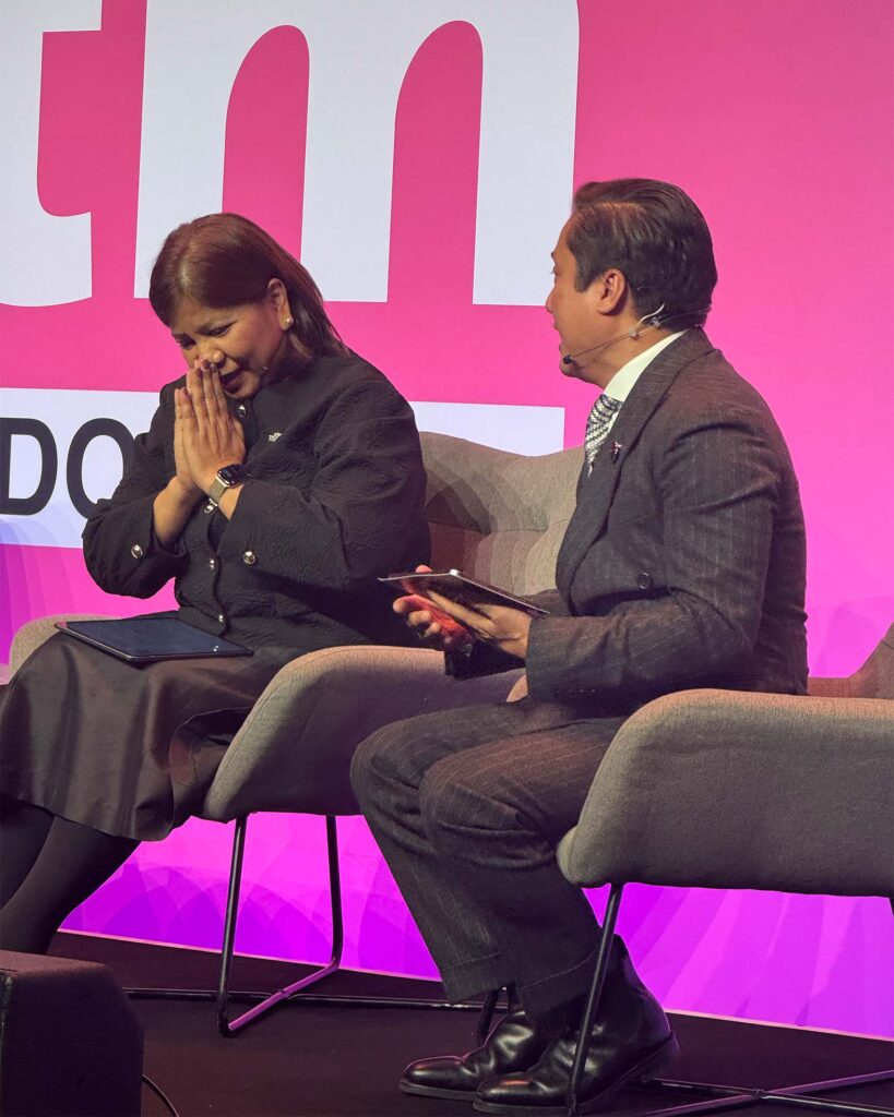 Two speakers sit in armchairs on the Orange Theatre stage. One holds their hands together in a traditional Thai greeting while the other looks on, smiling.