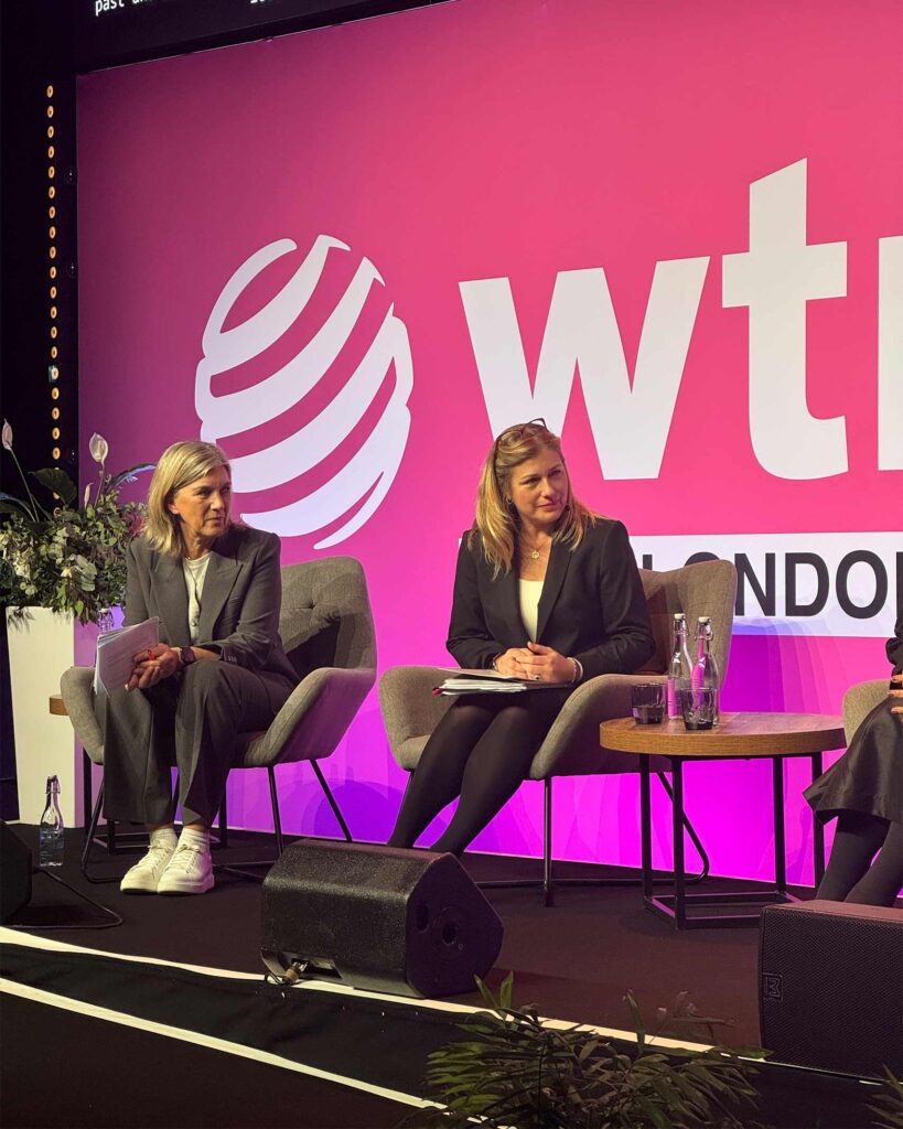 Two women sit on stage under the WTM London logo, listening attentively. Both wear business suits and have notes and water bottles in front of them.